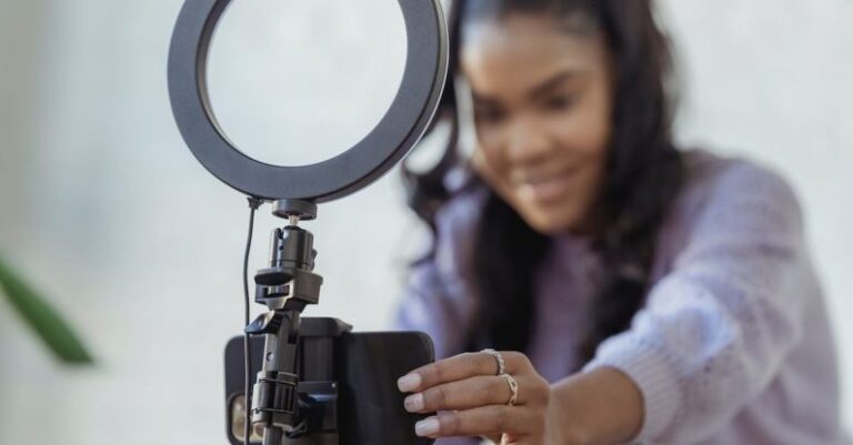 Video Content - Cheerful young African American female blogger in stylish sweater smiling while setting up camera of smartphone attached to tripod with ring light before recording vlog